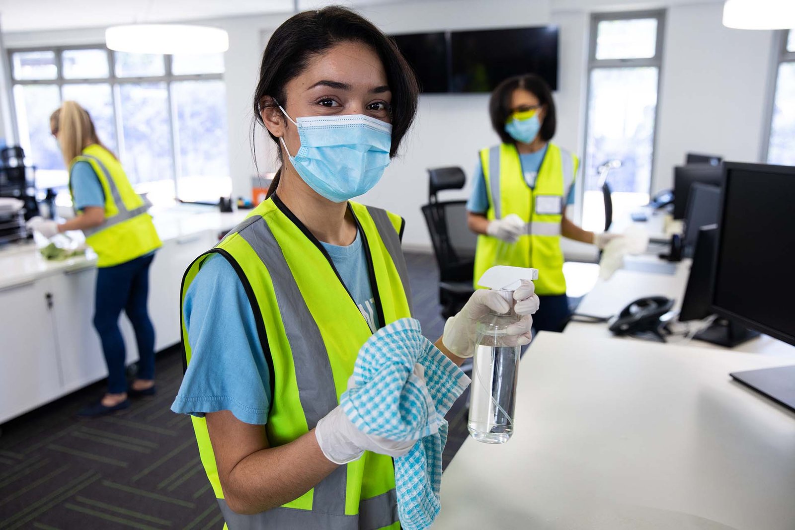 Portrait of mixed race woman wearing hi vis vest, gloves and face mask sanitizing office with disinfectant, colleagues in the background. Hygiene in workplace during Coronavirus Covid 19 pandemic.